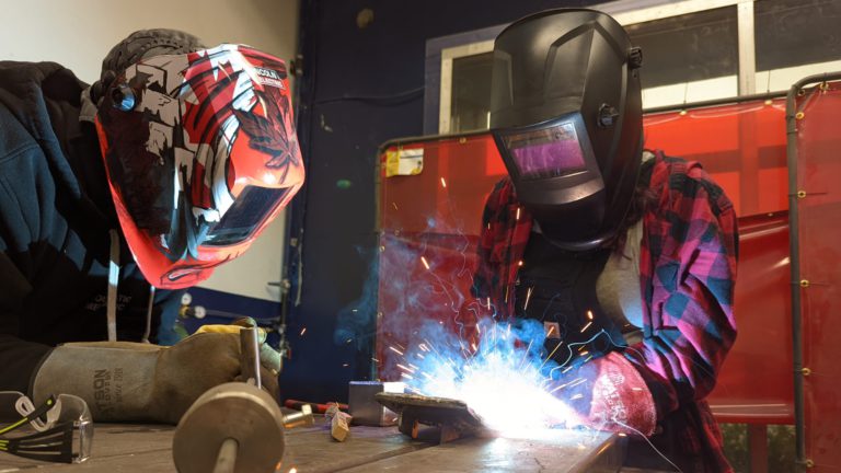 Student MIG welding with a Lincoln 210 multi-process welder at Maker Cube; helmet and gloves on with sparks visible, instructor Cory supervising in PPE (Langley, BC)