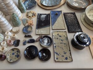 Table at Maker Cube in Langley, BC, covered with finished pottery pieces, class projects grouped in the center with additional member made work around the edges