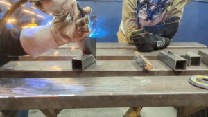 Instructor Corey at Maker Cube in Langley, BC, demonstrating an inside corner MIG weld on a rocket stove while a participant watches in a welding helmet, sparks flying