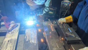 Participant MIG welding the side of a rocket stove at Maker Cube in Langley, BC, another student observing in a welding helmet, sparks visible, a second partially assembled stove on the table