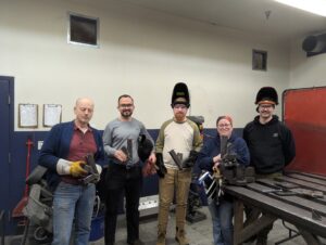 Group photo at Maker Cube in Langley, BC, instructor Corey with participants holding completed steel rocket stoves, smiling and a bit sweaty after class, welding PPE nearby