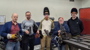 Group photo at Maker Cube in Langley, BC, instructor Corey with participants holding completed steel rocket stoves, smiling and a bit sweaty after class, welding PPE nearby