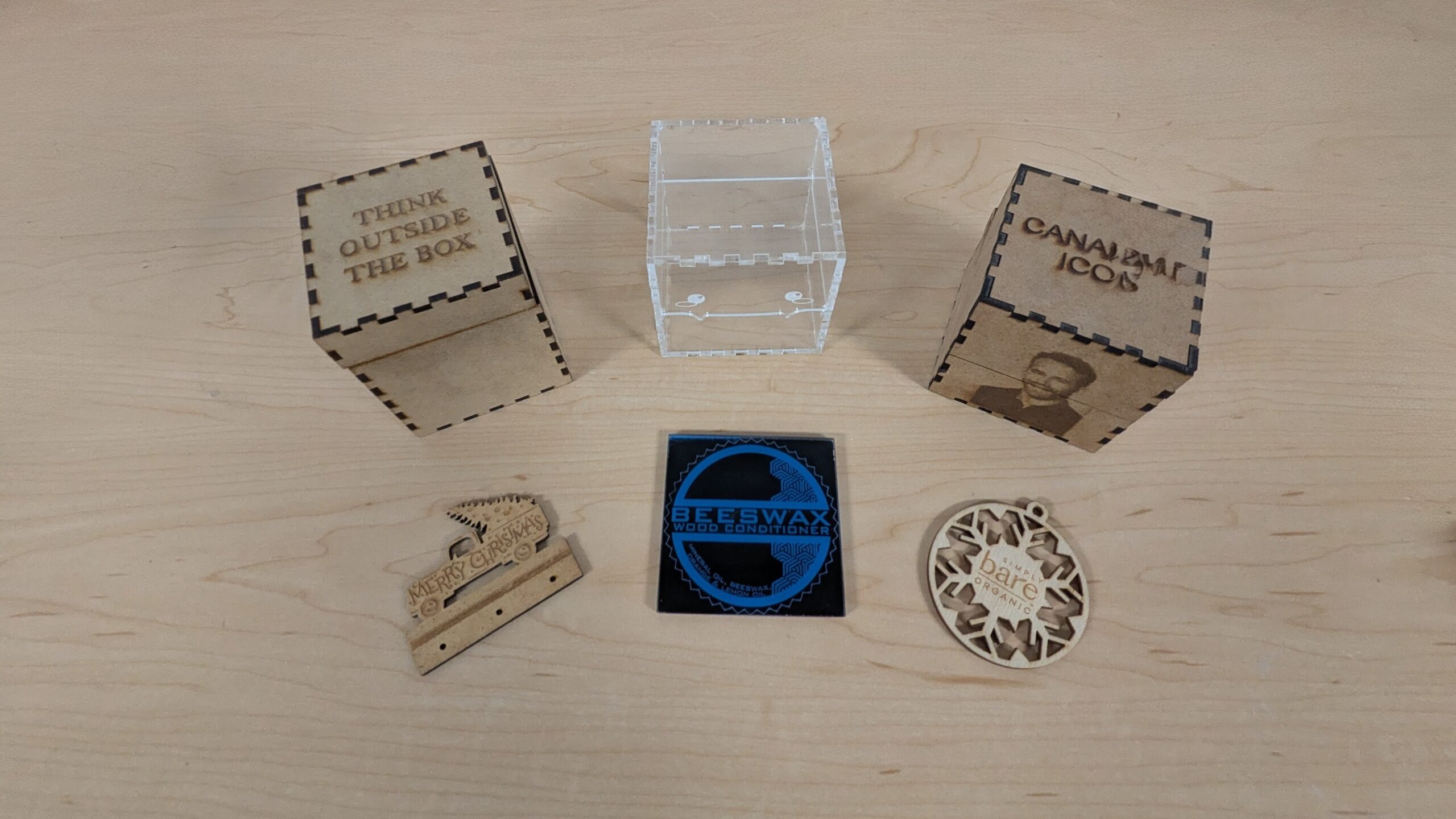 Three completed laser cut project boxes at Maker Cube, one in clear acrylic and two in MDF with different engraved designs, plus sample ornaments that show additional projects