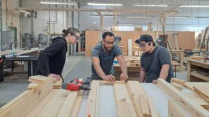 Instructor Adam at Maker Cube in Langley, BC, helping two members in the woodshop mark and measure lumber on a table with shop equipment in the background, prepping for planter boxes