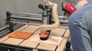 Process photo for the CNC Router Orientation in Langley, BC, serving Metro Vancouver and the Fraser Valley. A member observes and operates the router as it mills an Eastern maple pocket for a Padauk plug of a maple leaf. Class topics include workholding, bit selection, feeds and speeds concepts, setting X Y Z zero, running test passes, and pausing safely.