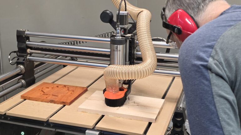Process photo for the CNC Router Orientation in Langley, BC, serving Metro Vancouver and the Fraser Valley. A member observes and operates the router as it mills an Eastern maple pocket for a Padauk plug of a maple leaf. Class topics include workholding, bit selection, feeds and speeds concepts, setting X Y Z zero, running test passes, and pausing safely.