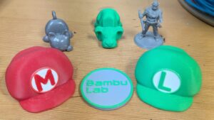 Assortment of 3D printed parts at Maker Cube in Langley, BC, including a Mario hat, a Luigi hat, two animal keychain phone holders, a tabletop miniature, and a token labeled “bambu labs”