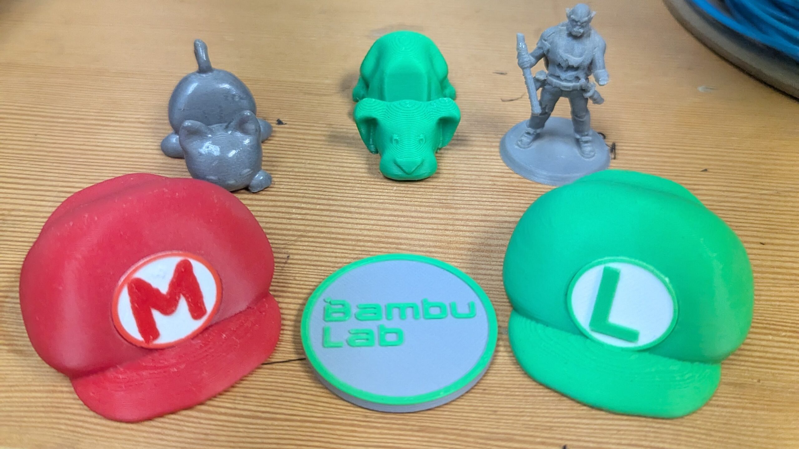 Assortment of 3D printed parts at Maker Cube in Langley, BC, including a Mario hat, a Luigi hat, two animal keychain phone holders, a tabletop miniature, and a token labeled “bambu labs”